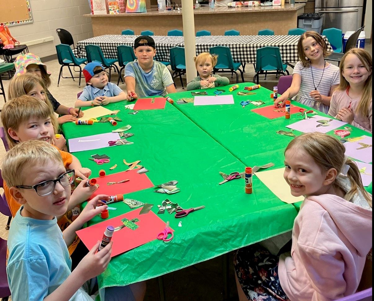 Ten kids sitting at a table with a green tablecloth, working on a collage craft project.